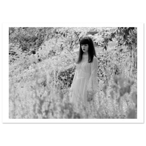 A solitary figure in a flowing white dress stands among overgrown ruins, suspended between light and shadow. Captured in timeless black and white, this photograph evokes quiet tension — innocence contrasted with something unspoken.